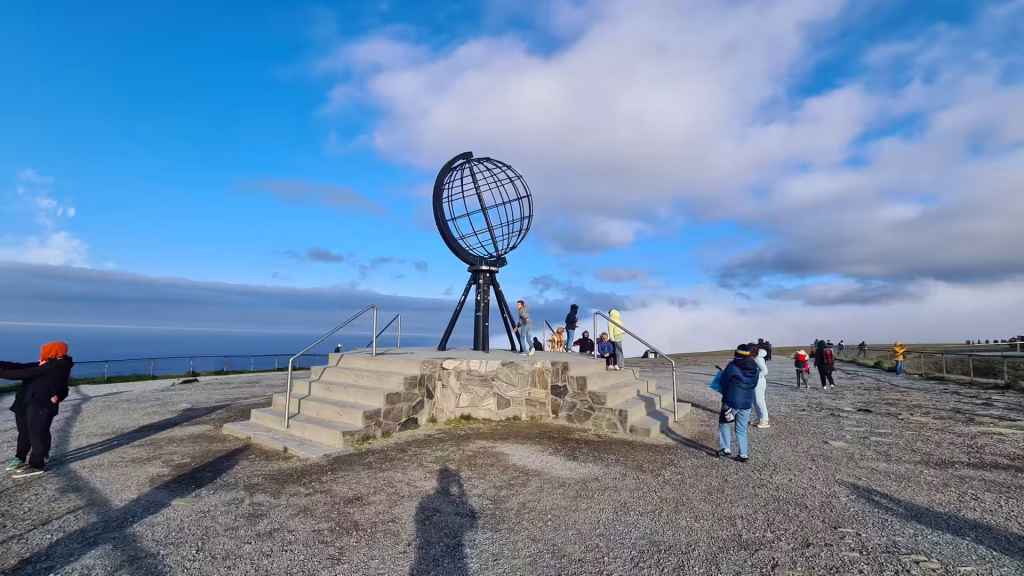 People gather on a gravel plateau around a large open framework globe sculpture on a stone pedestal, at the North Cape in Finnmark, Norway.