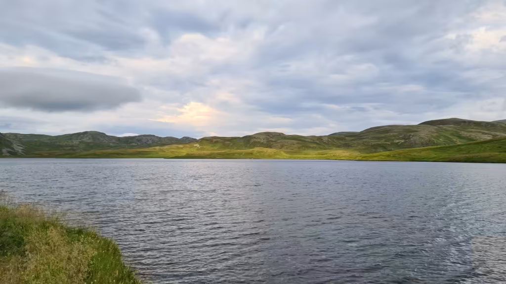 Grey rippled water stretches towards low green hills under a layered overcast sky, at Skarsvag on Mageroya in Finnmark, Norway.