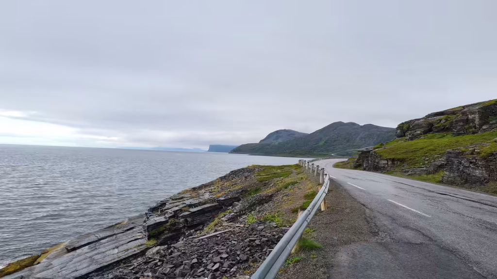A two lane road with a metal guardrail curves between a rocky shore and a steep cut of layered rock, in Porsanger in Finnmark, Norway.