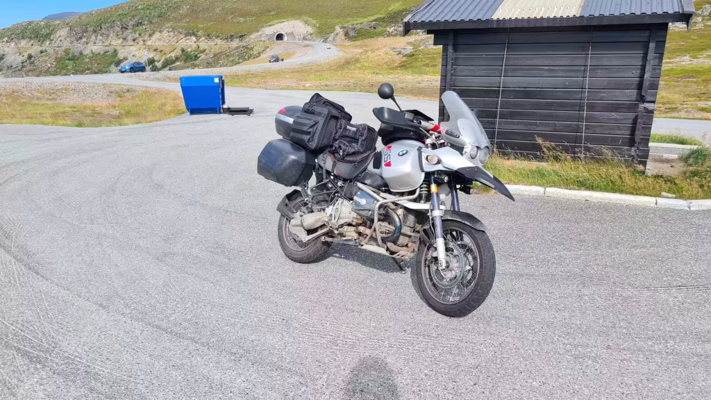 A loaded BMW GS adventure motorcycle rests on asphalt beside a small wooden hut with a road rising towards a tunnel mouth in the hill, at Hammerfest in Finnmark, Norway.