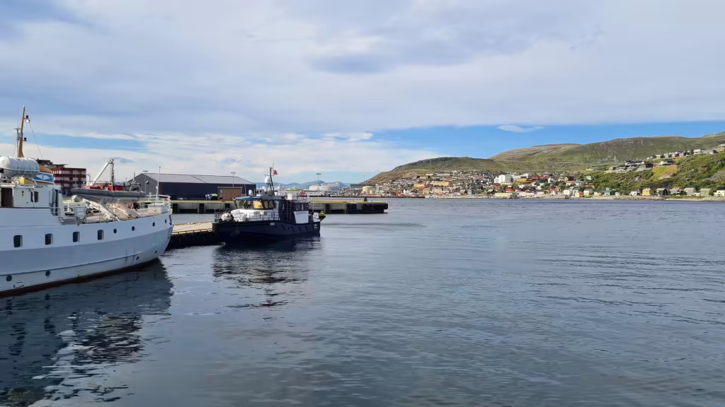 A large white vessel with rows of portholes and a small dark boat with a flag lie moored at a pier opposite a hillside town, at Hammerfest in Finnmark, Norway.