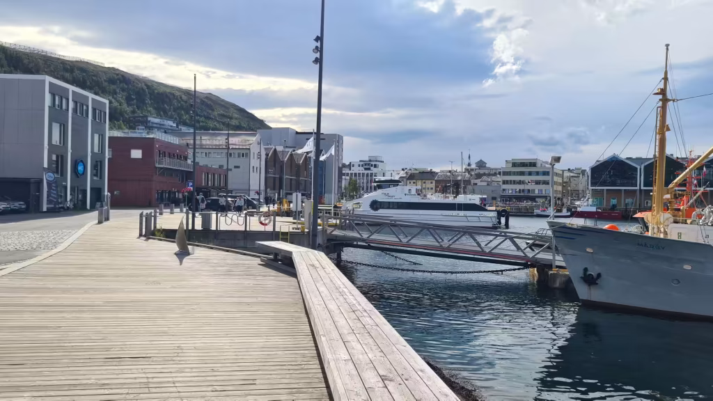A white catamaran ferry is moored at a plank pier beside a waterfront building while a grey vessel with yellow masts waits further out, at Hammerfest in Finnmark, Norway.