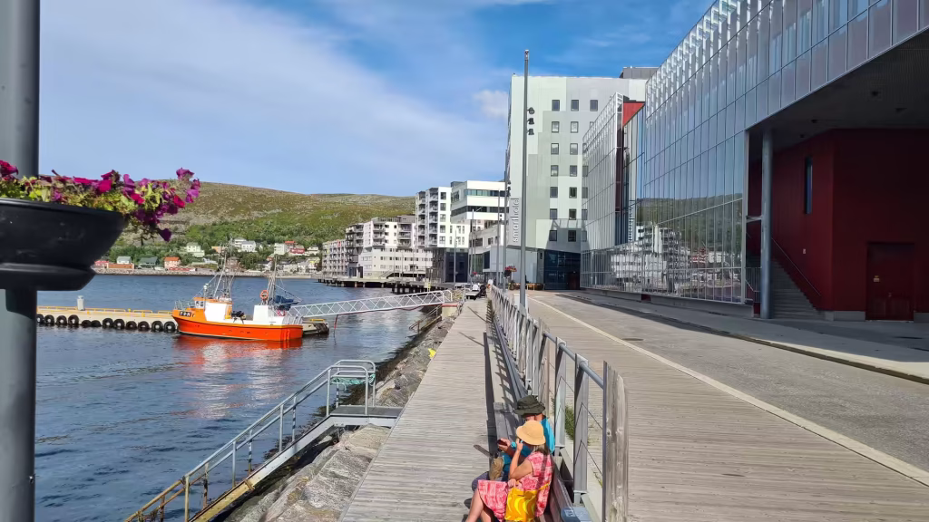 Two people sit on the boards of a waterside promenade beside flower planters while a bright orange boat lies docked at the pier below, at Hammerfest in Finnmark, Norway.