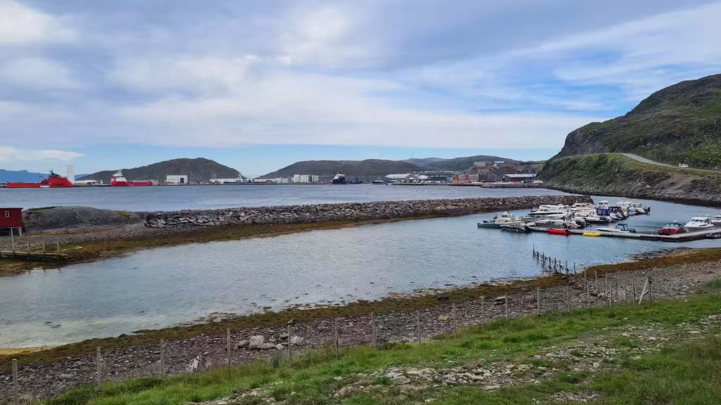 A long rock breakwater separates a shallow tidal inlet from a small boat marina below a steep green hill and a distant port, at Hammerfest in Finnmark, Norway.