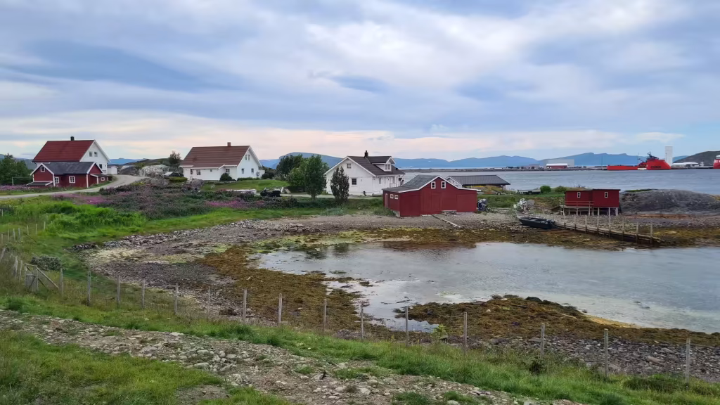 Red and white houses stand scattered along a rocky shoreline above a shallow tidal pool with distant mountains and a red vessel beyond, at Hammerfest in Finnmark, Norway.