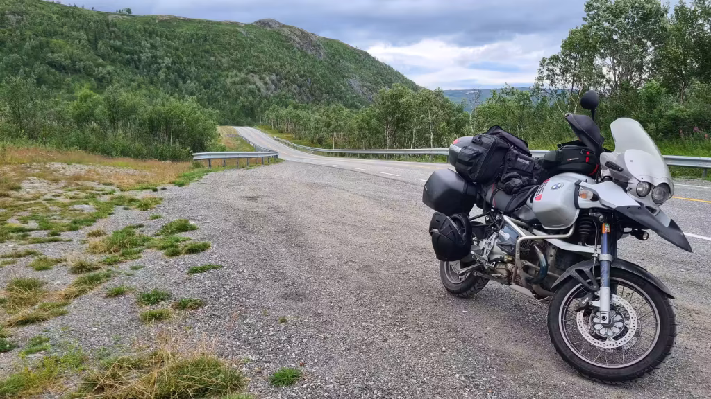 A loaded BMW GS adventure motorcycle stands on a gravel shoulder where a paved road curves uphill into green hills under an overcast sky, at Alta in Finnmark, Norway.