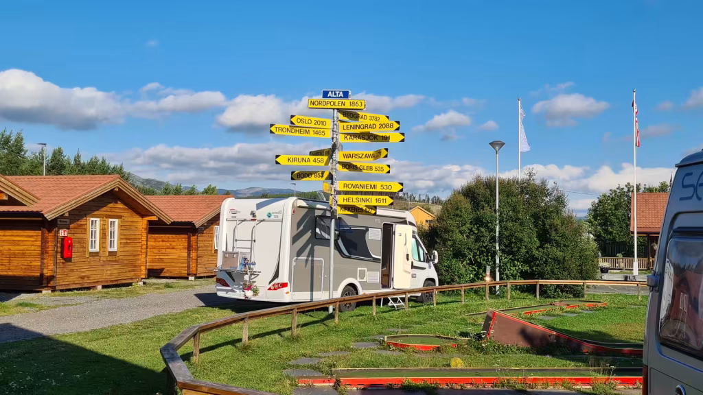 A white motorhome is parked on gravel beside a tall signpost of yellow distance signs near two wooden cabins and a miniature golf course, at Alta in Finnmark, Norway.