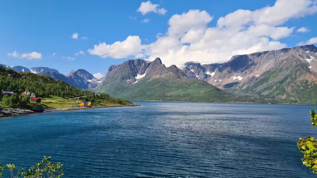 Dark rocky peaks carrying patches of snow rise across a wide blue fjord beside a few small red and yellow houses on the shore, at Oksfjord in Finnmark, Norway.