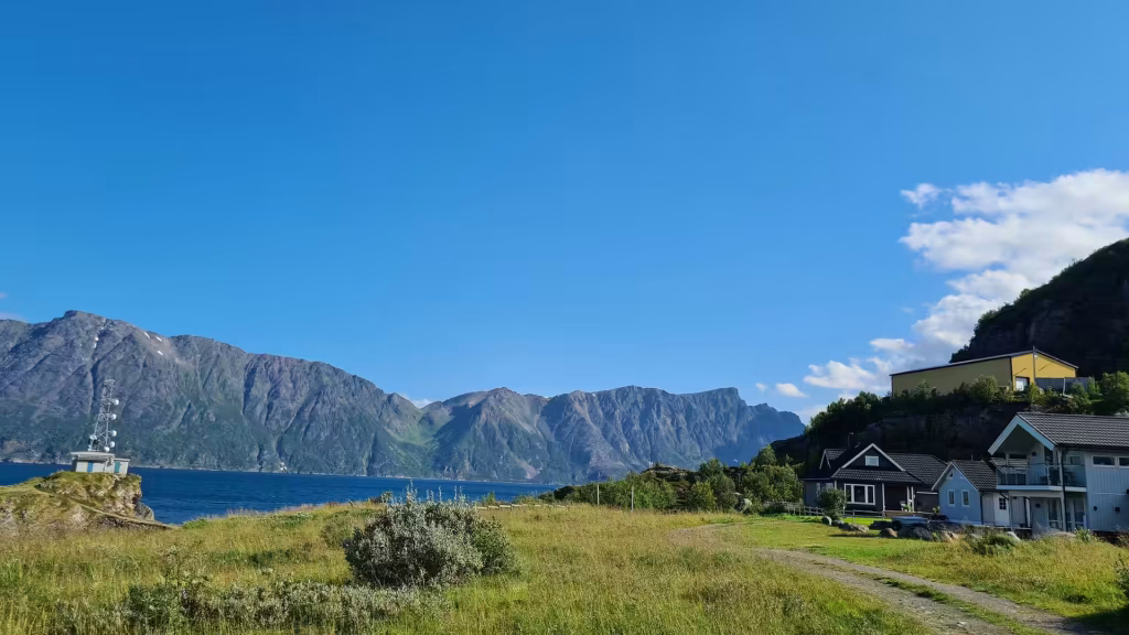 Steep grey mountains rise across a wide blue stretch of water beside a rocky point that carries a metal lattice tower, at Oksfjord in Finnmark, Norway.