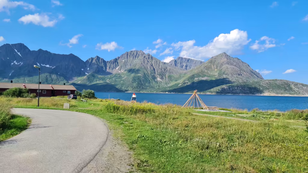 A grey road curves past a long brown building with a flag towards a conical frame of poles on the grassy shore, at Oksfjord in Finnmark, Norway.