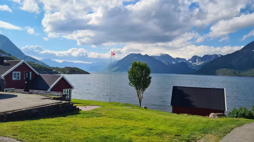 Red wooden buildings with white trim stand above a green lawn where a flag flies from a tall pole beside calm water, at Oksfjord in Finnmark, Norway.