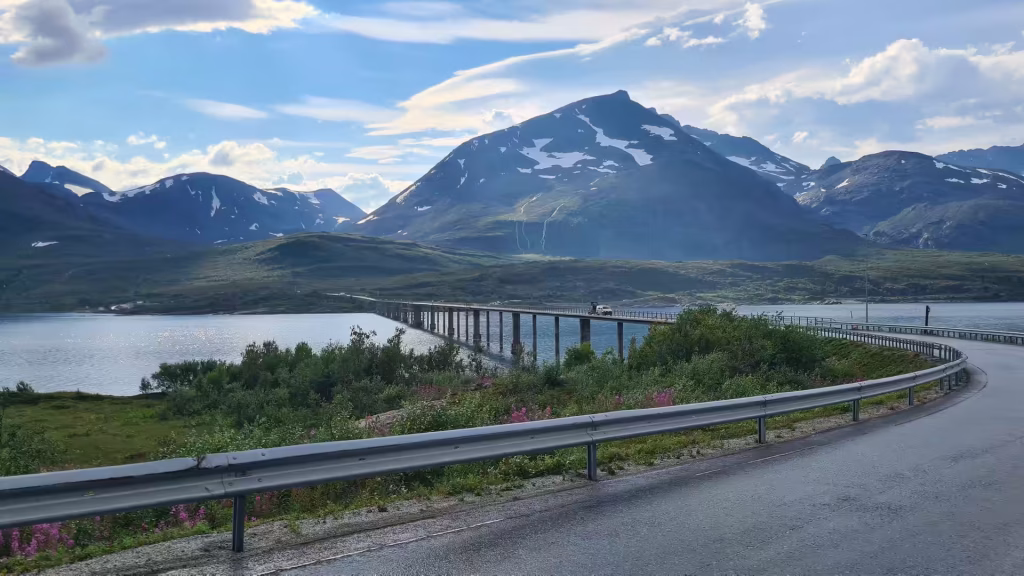 A curving road with a metal guardrail leads towards a long low bridge crossing water below a rugged mountain patched with snow, at Skjervoy in Troms, Norway.