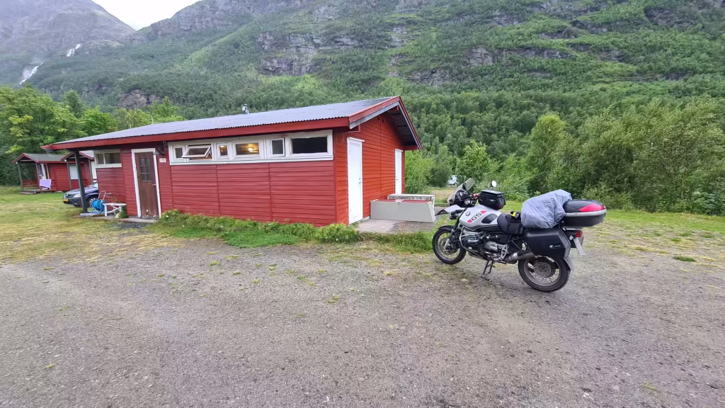 A loaded BMW GS adventure motorcycle stands on its centre stand before a long red timber building below steep green slopes, at Birtavarre in Troms, Norway.