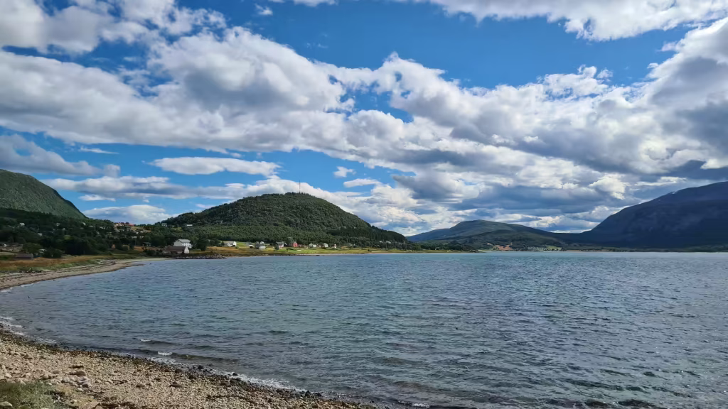 A gravel shoreline curves past calm water toward a wooded hill topped by a slim mast above green fields and scattered houses, at Tjeldsund in Norway.