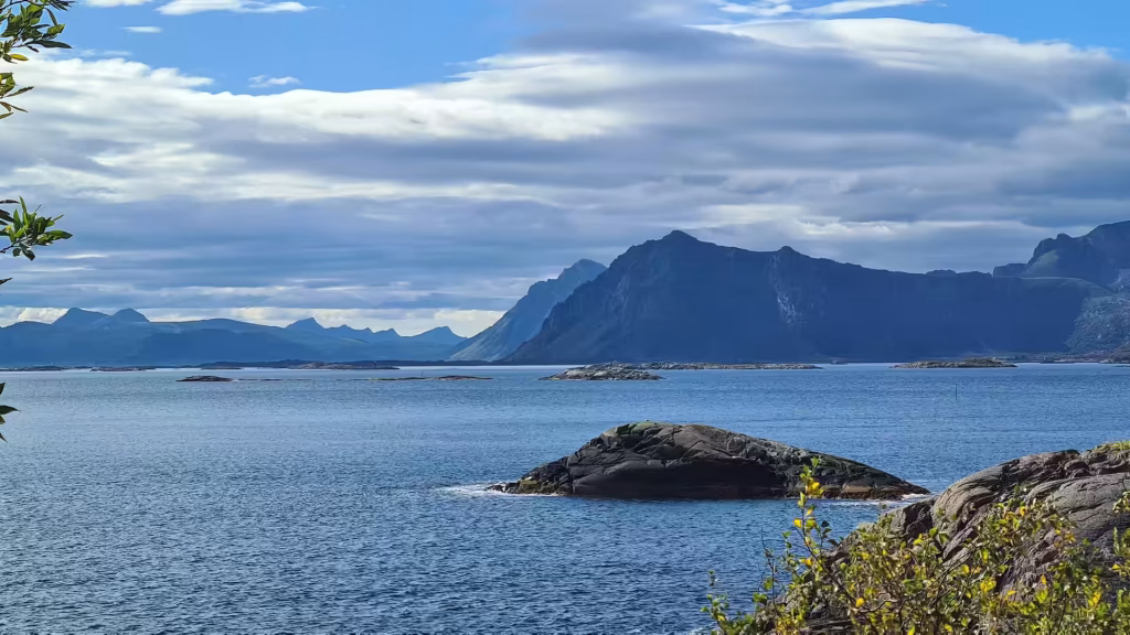 Dark rounded rocks sit in rippled blue water while a jagged mountain massif rises behind low skerries under soft grey cloud, in the Lofoten islands, Norway.