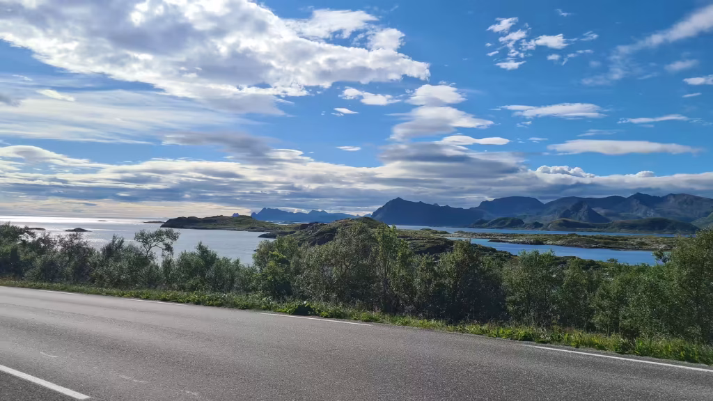 A grey asphalt road with white markings runs beside a band of shrubs above calm water dotted with small green islands, in the Lofoten islands, Norway.