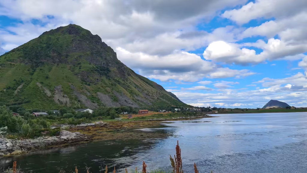 A steep green mountain rises above small red and white cabins on the shoreline of a calm body of water under a partly cloudy sky, in the Lofoten islands, Norway.