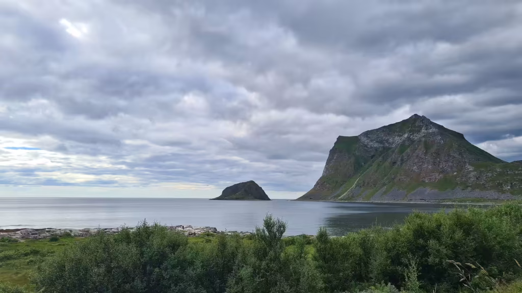 A small dark rocky islet sits in calm grey water below a green headland under a heavily overcast sky, in the Lofoten islands, Norway.