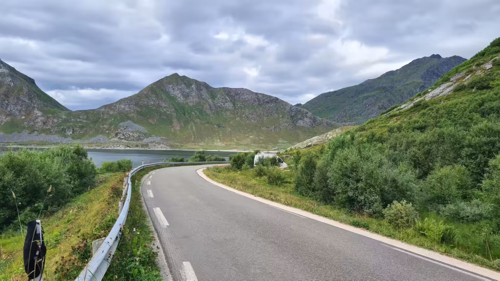 A paved road with a white centre line curves past a metal guardrail above a calm body of water below rugged mountains, in the Lofoten islands, Norway.