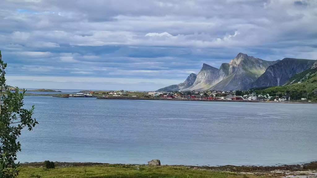 Small red and white buildings line the far shore of a wide calm bay below steep rugged mountains, at Ramberg on the Lofoten islands, Norway.