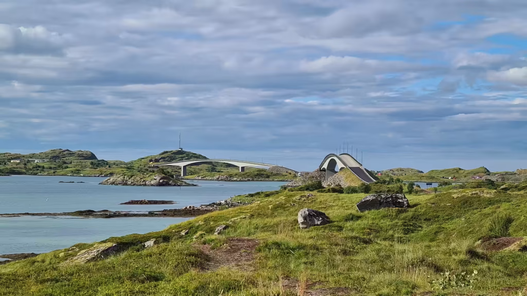 A low long bridge and a nearby arched bridge cross calm water between grassy rocky islets under a partly cloudy sky, at Ramberg on the Lofoten islands, Norway.