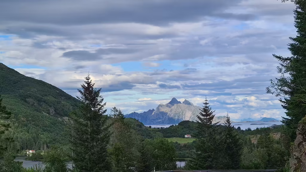 A jagged mountain ridge rises beyond a dark stretch of water, framed by conifer branches under a layered cloudy sky, at Leknes on the Lofoten islands, Norway.