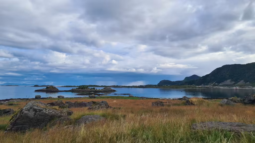Golden grass and large grey boulders line the water where small rocky islets sit below a steep dark hill under heavy cloud, at Stamsund on the Lofoten islands, Norway.