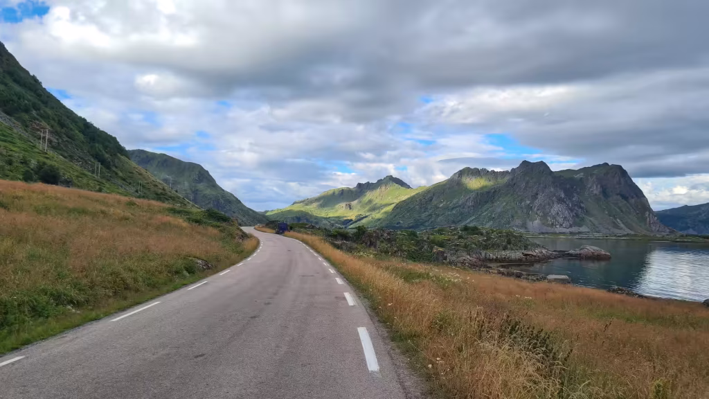 A grey road with white markings curves along a grassy hillside beside dark water, with a vehicle ahead and utility poles running uphill, at Stamsund on the Lofoten islands, Norway.