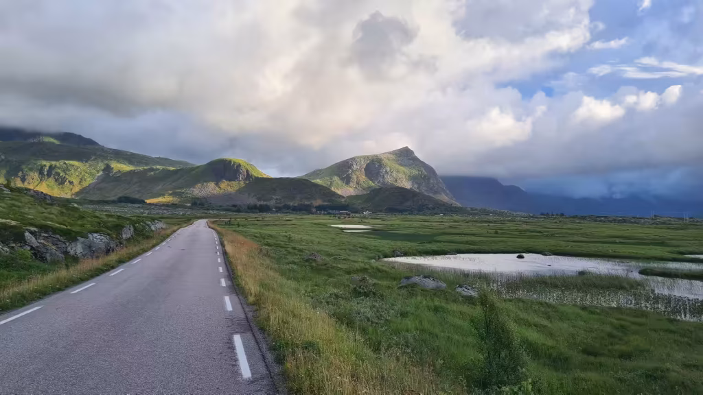A paved road with white lane markings runs past a flat green marsh whose shallow ponds mirror a bright cloudy sky below rocky hills, in the Lofoten islands, Norway.