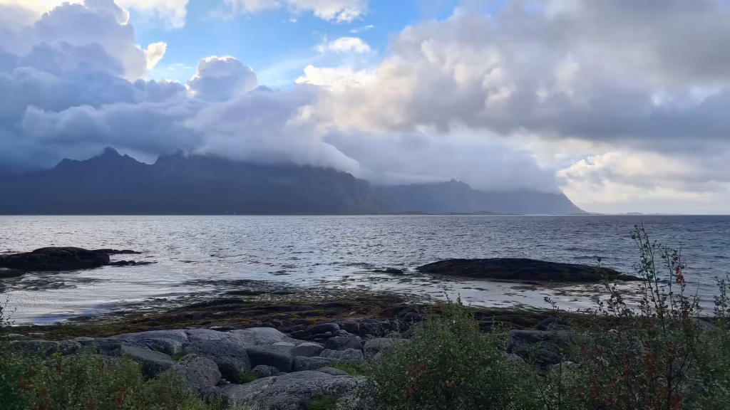 Grey boulders and low shrubs edge a wide expanse of rippled water where dark rocky islets sit below a cloud covered mountain range, in the Lofoten islands, Norway.