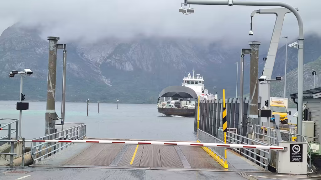 A ferry with a dark hull and white wheelhouse nears a metal ramp beside a lowered striped barrier at a wet terminal under low cloud, at Narvik in Norway.