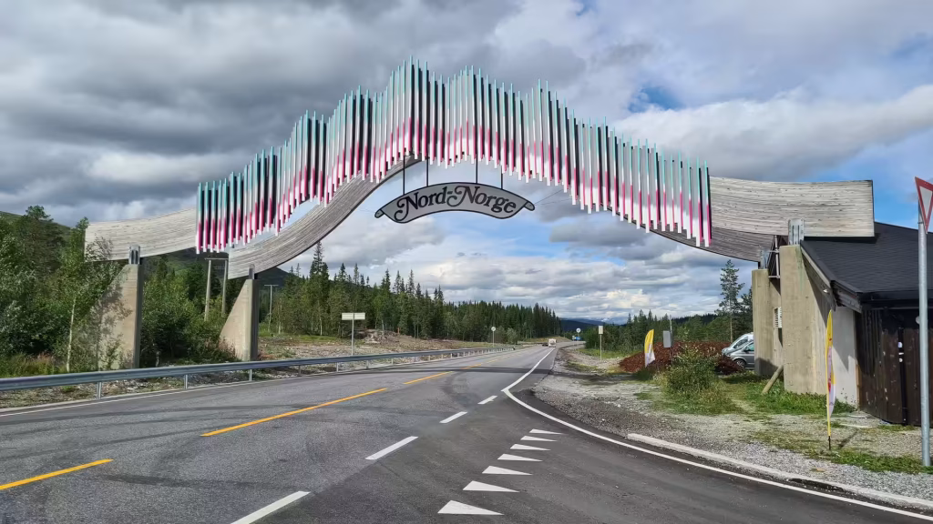A curved slatted arch carrying a scroll shaped sign spans a paved two lane road beside conifers and a small building, at Namsskogan in Norway.