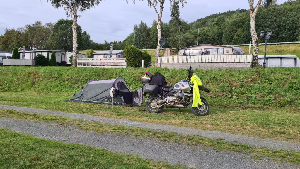 A loaded BMW GS adventure motorcycle stands beside a small grey tent on a grassy pitch lined with static caravans, on the Inderoy peninsula in Norway.