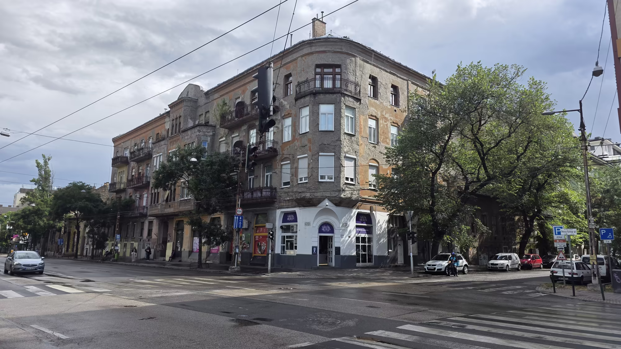 A weathered corner building with ground floor shops stands at a Budapest intersection with zebra crossings, parked cars and overhead tram wires under an overcast sky.